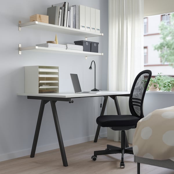 Modern minimalist office setup: white TROTTEN desk, black mesh chair, laptop, and lamp. Large window with curtains.