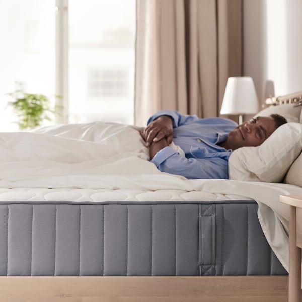 A person sleeps comfortably on a VÅGSTRANDA mattress in a cosy bedroom, with soft lighting and a window in the background.