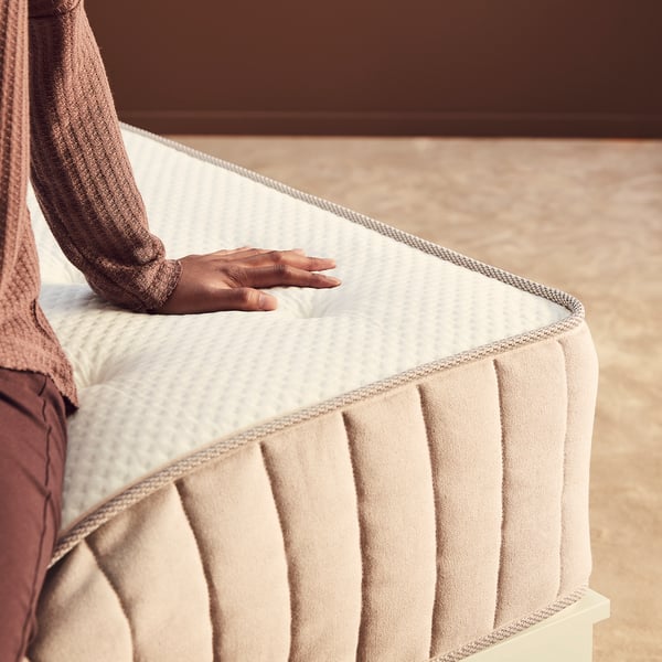 A person pressing a beige foam mattress, highlighting its layers and texture.