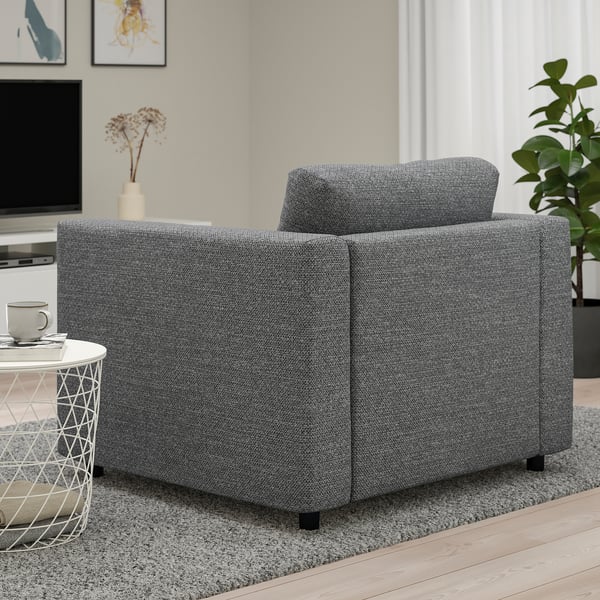 A modern grey fabric armchair in a living space with a side table and mugs, tv, and plants.