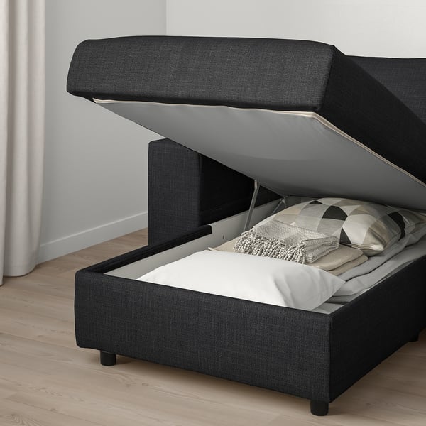 Black sofa with raised ottoman revealing storage. Inside, white blanket and pillows visible. Floor is light wood, walls white.