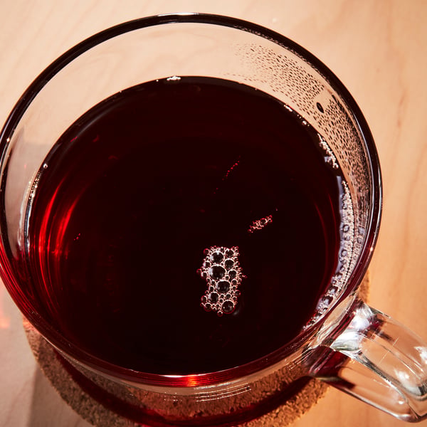 Clear glass mug filled with dark red, spiced beverage. Small bubbles visible, suggesting warmth. Placed on a wooden table.