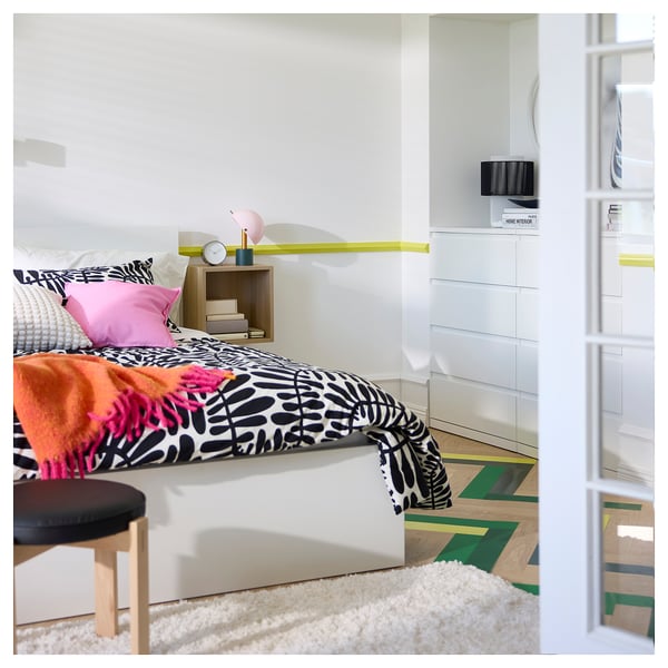 Bright bedroom with white bed, colourful pillows, patterned bedding, wooden stool, bedside table, and a fluffy white rug on wooden floors.