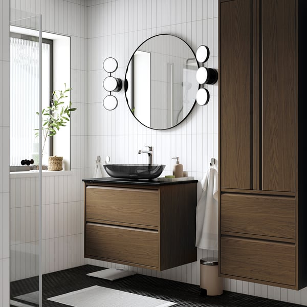 Meuble de salle de bain moderne en bois avec lavabo en verre, miroir, lumières, serviette et plante.
