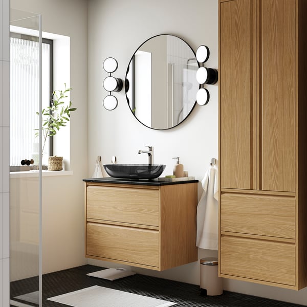 Salle de bain moderne avec meuble en chêne ÄNGSJÖN, lavabo sombre. Miroir rond, serviettes, plante.