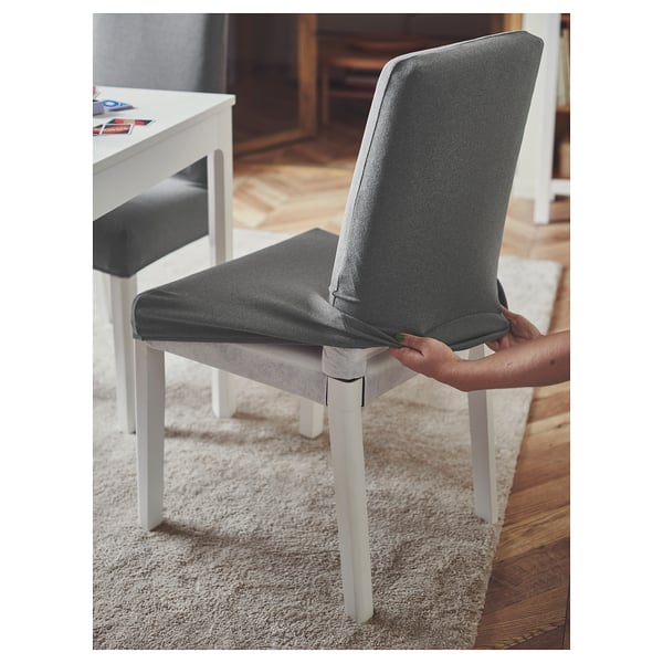 Une personne pose une housse de chaise ÄSPHULT en polyester gris sur une chaise blanche.