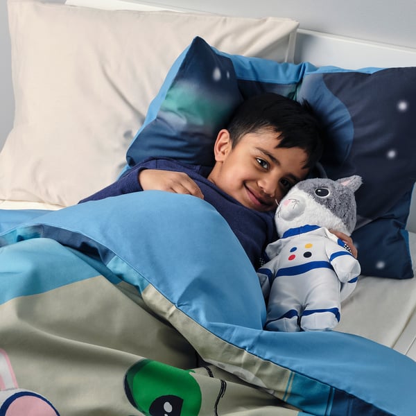 Garçon au lit avec chat gris en peluche en costume bleu, draps galaxie.