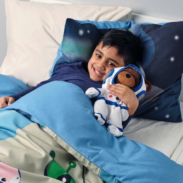 Garçon allongé sur une literie bleue tenant un ours en peluche astronaute, souriant joyeusement.