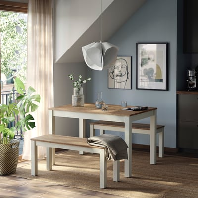 Salle à manger moderne avec banc marron clair ÅLHULT, jeté gris, vase de fleurs blanches et art abstrait sur mur bleu clair.