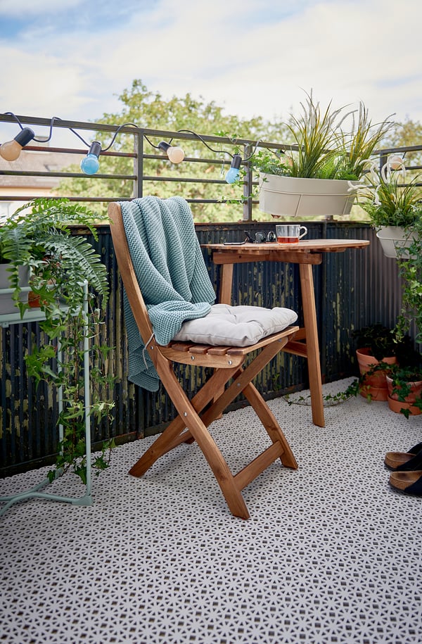 Configuration de balcon confortable avec une terrasse en plastique blanc ALTAPPEN, une chaise pliante en bois, des coussins, une table, des lampes suspendues et des plantes en pot.