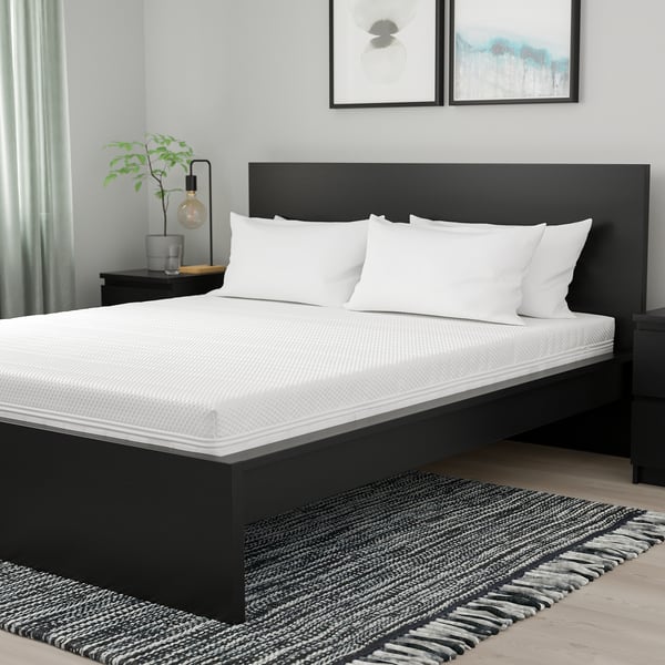 Chambre moderne avec matelas noir, literie blanche, plante, art, tapis sombre.
