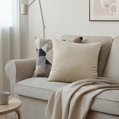 Le coussin en velours côtelé beige ÅSVEIG sur un canapé beige. Il est de forme carrée avec des lignes verticales, de taille moyenne entre autres.