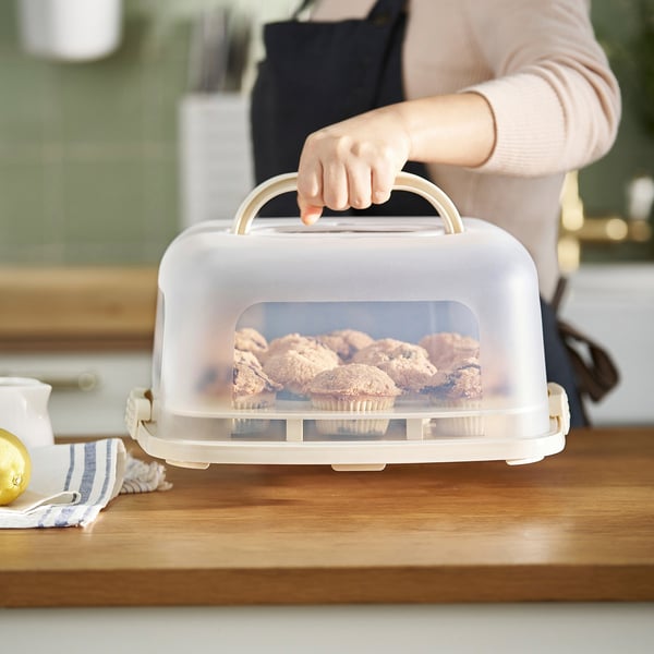 Une personne tenant un porte-gâteau avec des cupcakes. Le transporteur possède un couvercle et une poignée pour faciliter le transport.