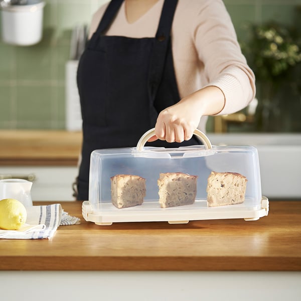 Personne tenant un porte-gâteau transparent avec couvercle, contenant trois parts de gâteau sur une table en bois.