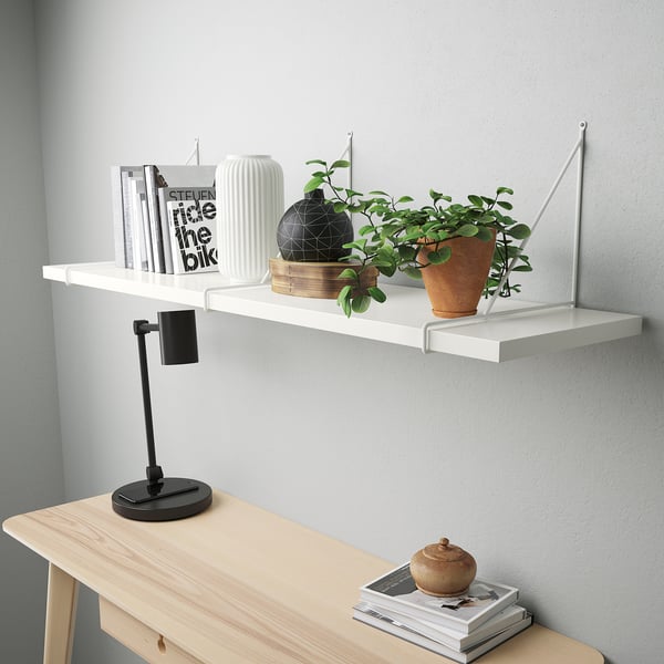 Étagère murale blanche moderne avec bord biseauté, contenant des livres, une plante en pot, un vase noir. En dessous, une console en bois clair avec lampe et magazines.