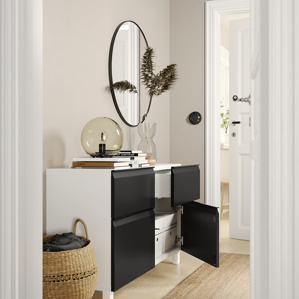 Entrée moderne avec buffet BESTÅ blanc et noir, portes ouvertes révélant des rangements, surmonté d’un décor, d’un miroir rond et d’un panier tressé.