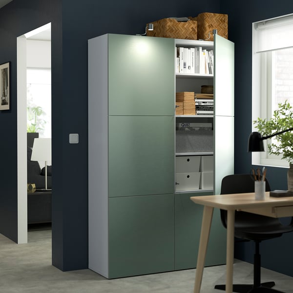 Bureau moderne avec unité de rangement verte BESTÅ, étagères réglables et lumières intégrées. Peut contenir des livres et des fournitures, associés à un bureau et une chaise en bois.