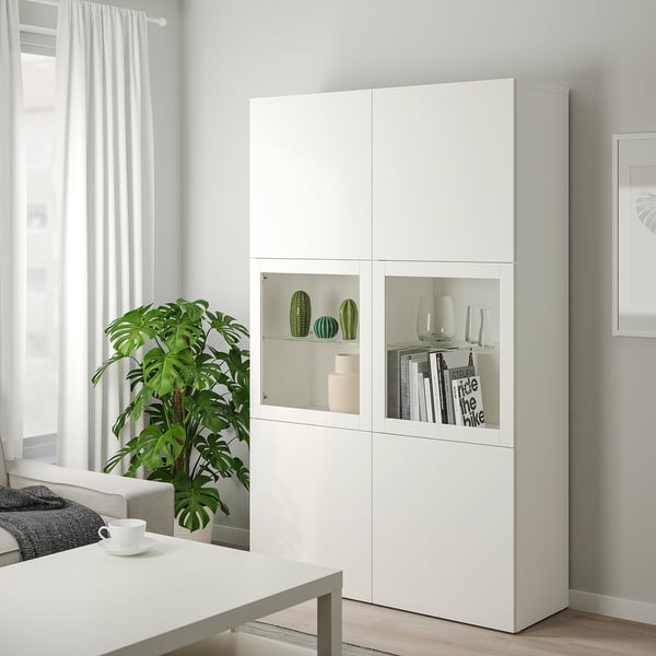 Grande armoire BESTÅ blanche avec portes en verre, pour ranger des objets décoratifs. Un design moderne et minimaliste, placé à côté d’une grande plante.
