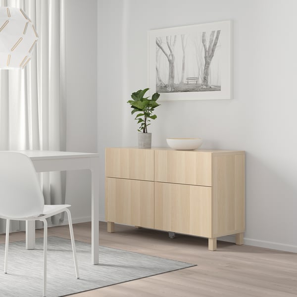 Chambre minimaliste avec armoire en chêne BESTÅ contre mur blanc, avec portes, plante, bol et image d’arbres encadrés près du bureau et de la chaise.