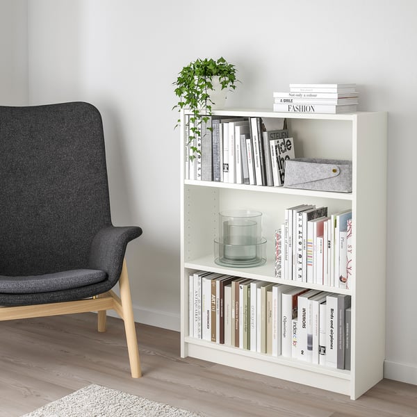 Étagère BILLY blanche avec 3 étagères, contenant des livres, une bougie et une plante. Chaise grise à côté, s’insère parfaitement dans le coin.