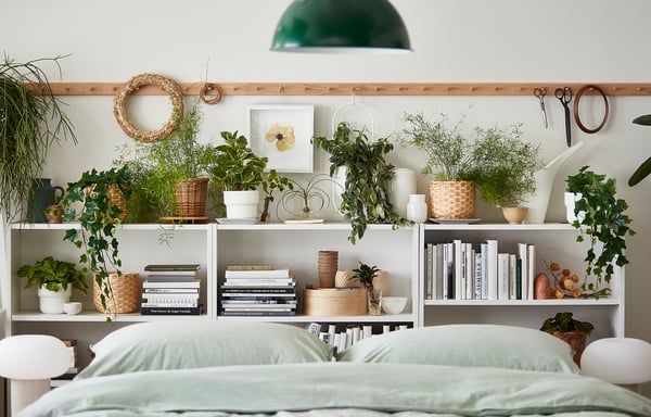 Chevet : étagère BILLY blanche avec des plantes vertes, des livres et un décor de panier en osier. Literie verte en dessous.