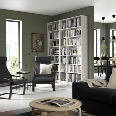 Étagère blanche modulaire remplie de livres, entre des murs verts, avec des meubles noirs, créant un coin lecture confortable.