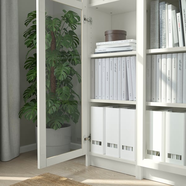 Bibliothèque BILLY blanche fermée avec portes vitrées, permettant de ranger des livres et une plante en pot, reflétant la lumière naturelle.