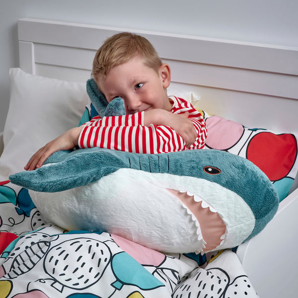 Un jeune garçon câline un requin bleu en peluche assis sur un lit. Le requin est grand et doux, parfait pour les câlins et les jeux.