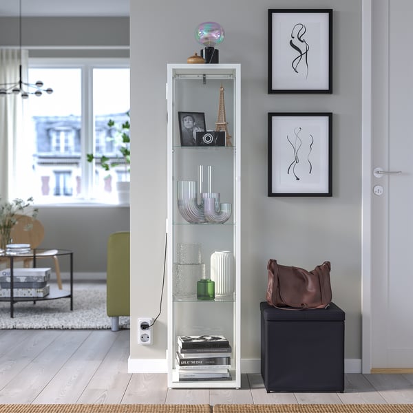 Salon moderne avec armoire blanche BLÅLIDEN, étagères décorées, cadres d’art et pouf sombre.