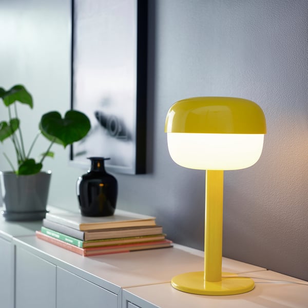 Lampe BLÅSVERK jaune vif sur table, entourée de livres, de plantes et d’un vase ; Rétro, forme douce, design moderne, petite taille.