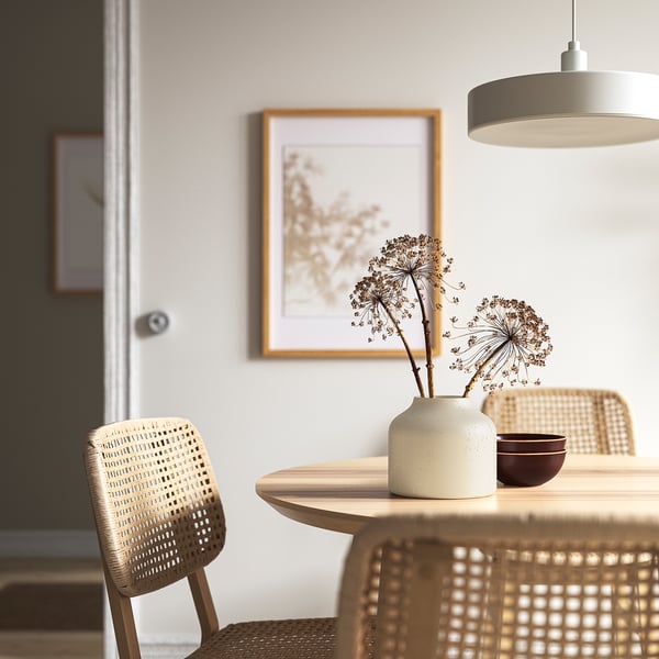 Salle à manger minimaliste avec table ronde, chaises en osier, vase, bol, suspension, imprimé nature.