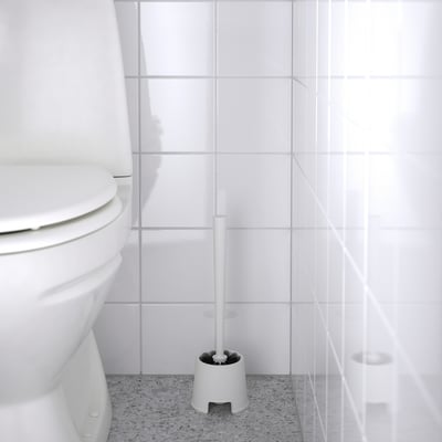 Une brosse de toilette blanche, BOLMEN, se trouve à côté d’une toilette sur un sol tacheté, prête à être nettoyée.
