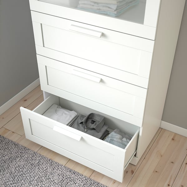 Commode blanche avec dessus en verre, un tiroir ouvert laissant apparaître des vêtements pliés. Ancré pour plus de sécurité.