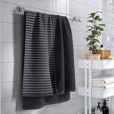 Une salle de bain moderne avec une serviette grise sur une barre chromée, à côté d’une étagère contenant des serviettes pliées, une plante et des articles de toilette.