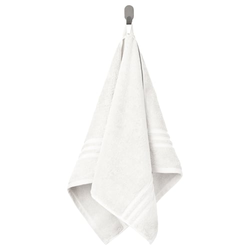 La serviette blanche BROKGLIM s’accroche soigneusement à un crochet, mettant en valeur son tissu doux et absorbant avec une bordure tissée décorative.