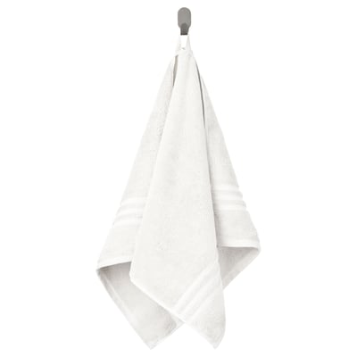 La serviette blanche BROKGLIM s’accroche soigneusement à un crochet, mettant en valeur son tissu doux et absorbant avec une bordure tissée décorative.