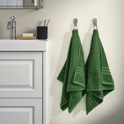 Serviettes bio BROKGLIM dans la salle de bain. Vert foncé, 100 % coton, plié sur des crochets argentés à côté de l’évier. Doux, absorbant, avec bordure tissée.