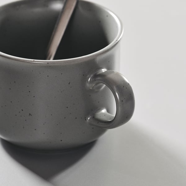 Tasse en grès, avec des mouchetures noires et une cuillère métallique à l’intérieur.