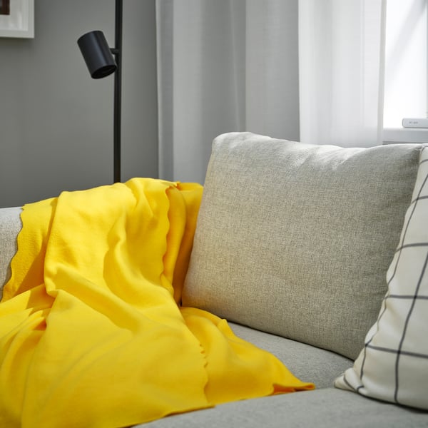 Jeté en polaire DAGGKÅPOR jaune vif sur canapé gris, doux, rectangulaire, à côté d’un grand oreiller blanc.