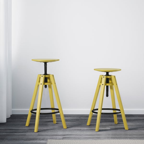 Deux tabourets de bar réglables jaunes, adaptés aux hauteurs de 90 à 110 cm, posés contre un mur blanc sur un sol gris.