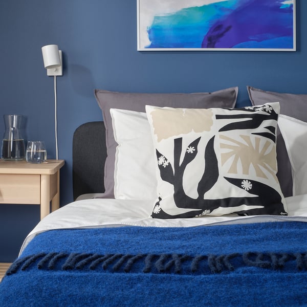 Chambre bleue avec lit fleuri, oreillers gris, jeté bleu, table de chevet en bois, carafe en verre, art.