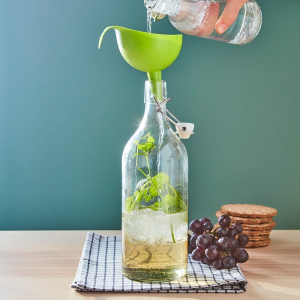 La main verse du liquide dans une bouteille avec des herbes à l’aide d’un entonnoir. Bouteille sur tissu à carreaux avec des raisins et des biscuits sur une table.