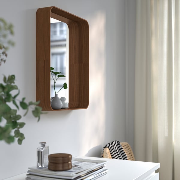 Miroir mural avec étagère, fenêtre réfléchissante. Tient un petit vase avec des plantes sur une étagère. Entouré de meubles blancs et de verdure.