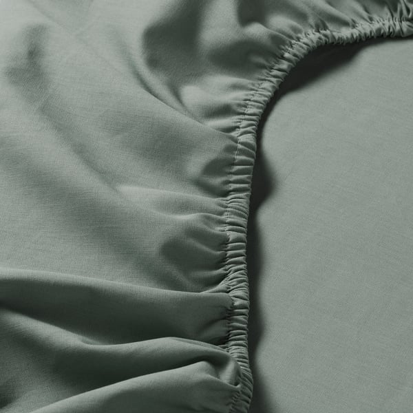 Gros plan du drap-housse DVALA en gris-vert, présentant une texture de coton lisse et un ourlet élastique, mettant l’accent sur la qualité et l’artisanat.