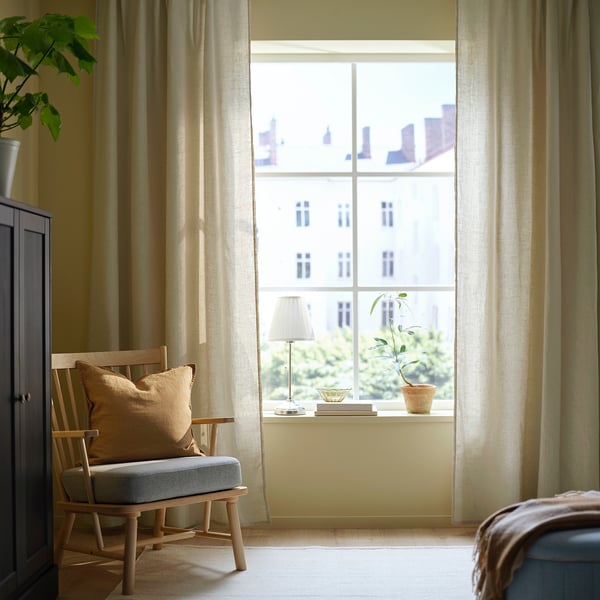 Chambre lumineuse avec de grands rideaux beiges DYTÅG, une chaise en bois, un coussin beige moelleux et une plante en pot sur le rebord de la fenêtre.