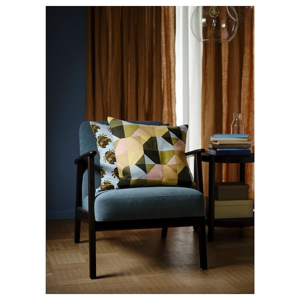 Des coussins graphiques modernes sur une chaise grise de style rétro avec des pieds en bois, positionnée près d’une petite table et d’une grande fenêtre avec des rideaux marron.