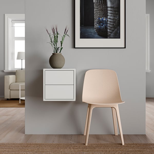 Unité de rangement EKET blanche minimaliste avec deux tiroirs, avec une chaise beige et une plante en pot sur le dessus. Mur gris avec art encadré. Parquet clair.