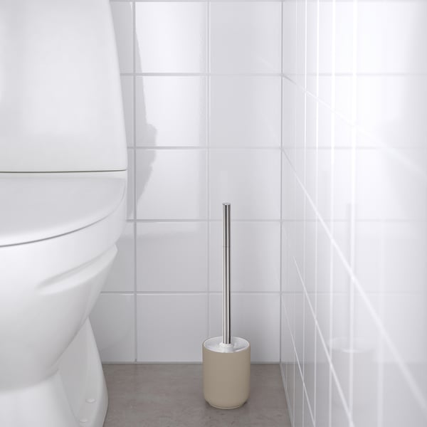 Salle de bain avec toilettes blanches et porte-brosse beige tenant une brosse métallique.