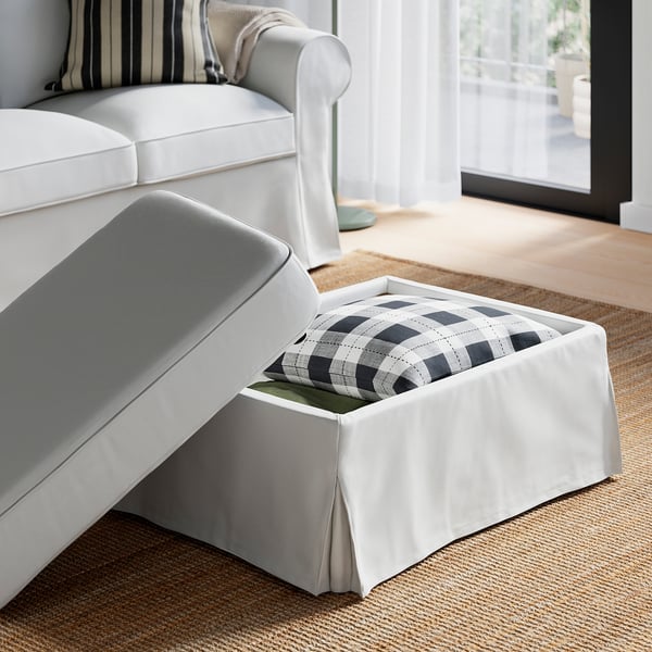 Canapé blanc avec ottoman, un côté ouvert montrant une couverture à carreaux noir et blanc à l’intérieur, posée sur un tapis texturé près d’une grande fenêtre.