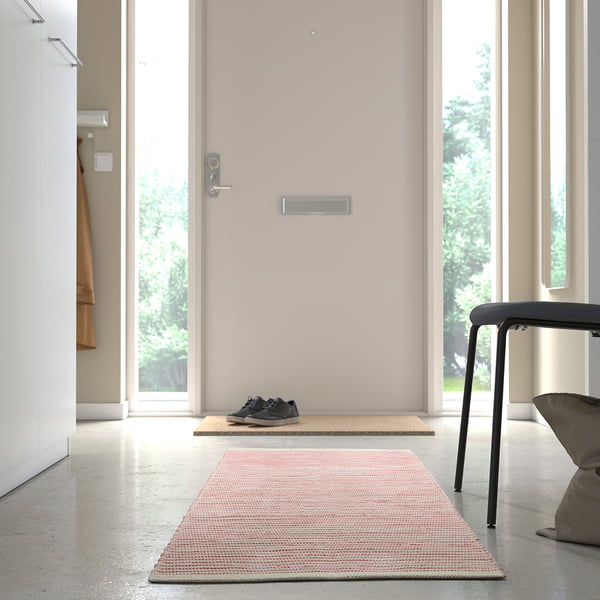 Une entrée minimaliste avec un petit tapis sur le sol près de la porte, et un autre tapis légèrement plus grand plus loin. Un tabouret et un cintre sont visibles.